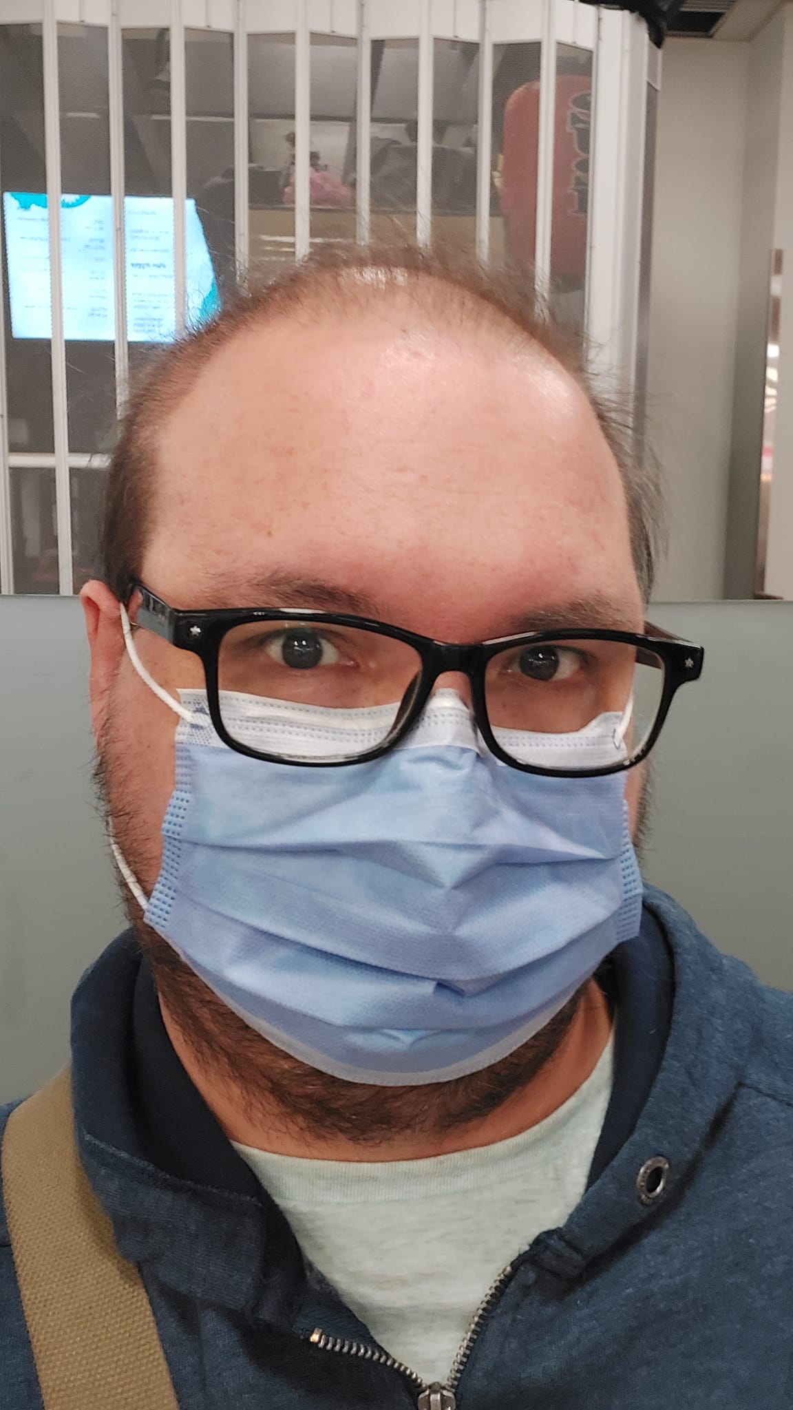 A man sits in a food court inside the hospital, wearing a sweater and surgical mask